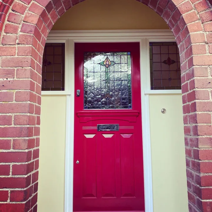 Crimson 1930s-style timber front door with a leaded stained glass panel, set in an arched brick porch with matching side panels