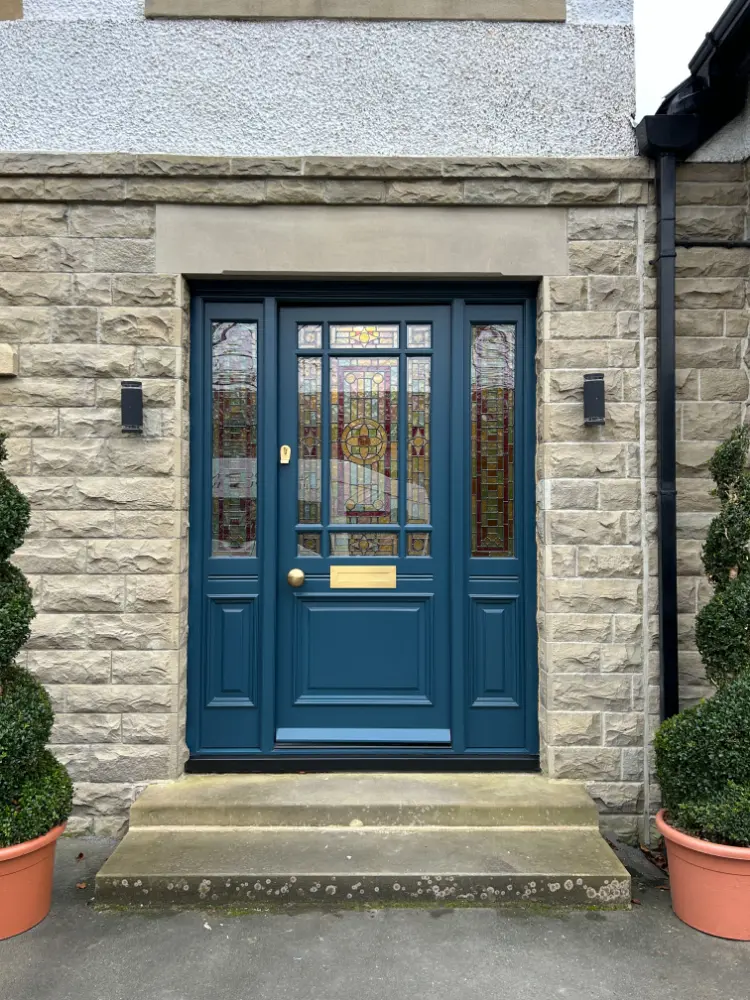 Blue painted timber front door with glazed upper panels and brass furniture on a stone-faced house, flanked by topiary in pots