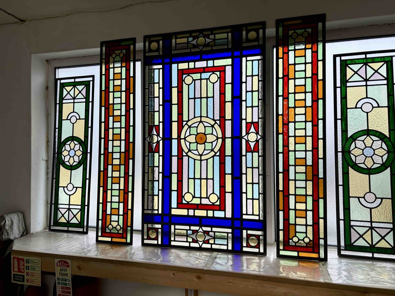 Elegant white double casement window with intricate stained glass panels featuring red and green floral motifs, brass handles, and decorative moulding.