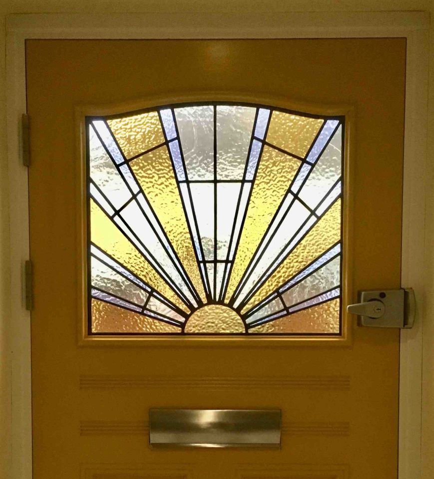 Traditional teal front door with decorative stained glass panels featuring geometric patterns, set in a red brick wall with a black house number plaque.