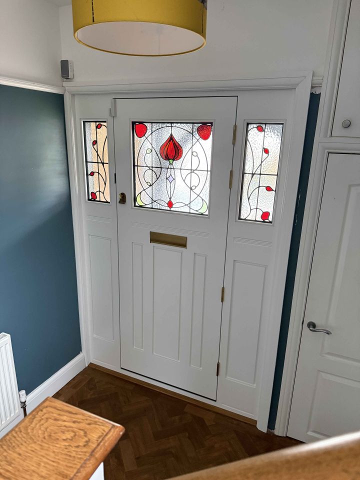 Blue front door with an intricate stained glass panel and matching side lights, set within a stone facade, flanked by topiary trees in terracotta pots.
