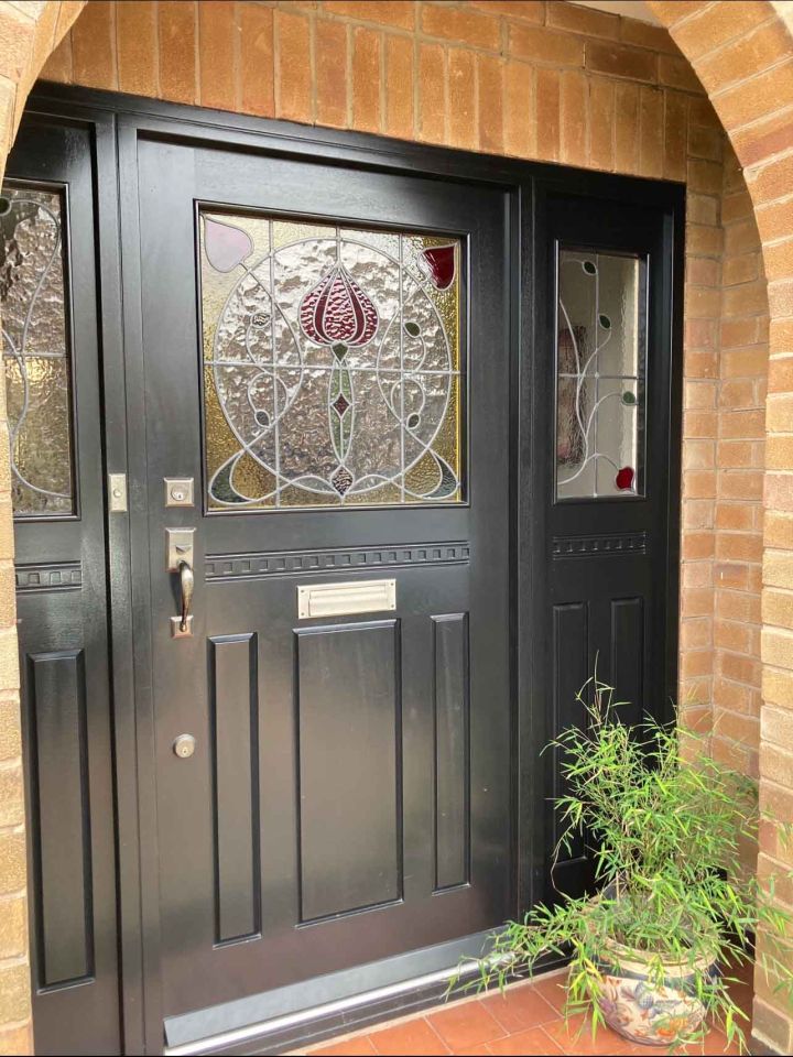 Black front door with an ornate arched stained glass panel featuring floral designs, flanked by matching side panels with decorative glass, set within a brick facade.